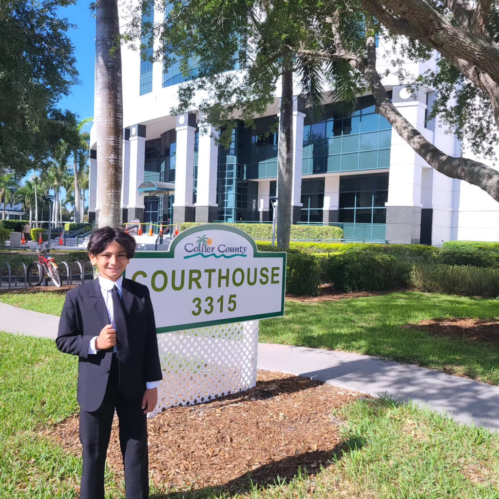 Esteban, son of José O. Castañeda, Jr., beside the Collier County Courthouse sign in Naples, Florida, during a bring-your-child-to-work visit.