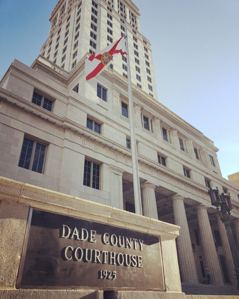 A warm-toned view of the historic Miami-Dade County Courthouse facade, plaque and Florida state flag in downtown Miami.